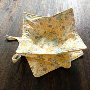 May include: A yellow and green floral patterned fabric bowl cozy. The cozy is folded in half and has two handles.