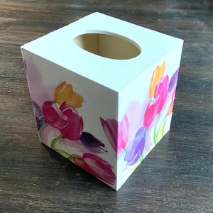 May include: White square tissue box with a watercolor floral design featuring pink, purple, and yellow tulips. The box has an oval opening on top for tissue dispensing. The floral pattern wraps around the sides of the box.