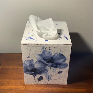 May include: A white tissue box with blue floral artwork and blue paint splatters. The box has a rectangular shape with a tissue emerging from the top. The floral design features large blue flowers.