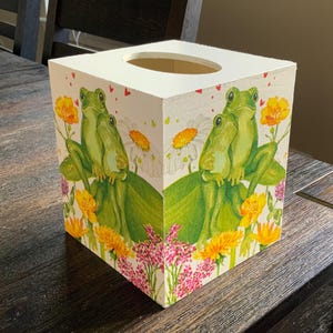 May include: A square tissue box with a painted design featuring two green frogs on a lily pad, surrounded by yellow and pink flowers, and white daisies. The box is white with a circular opening on top.