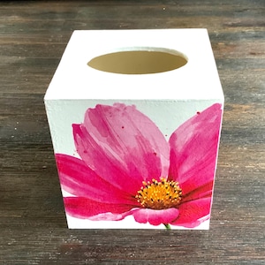May include: White square tissue box featuring a large, vibrant pink watercolor flower with a yellow center. The box has a smooth finish and a circular opening on top for tissue dispensing. The floral design adds a decorative touch.