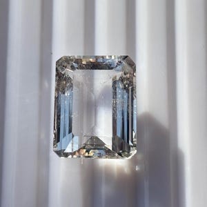 Könnte beinhalten: Ein klarer, rechteckiger Edelstein mit Smaragdschliff. Der Edelstein hat ein transparentes Aussehen mit inneren Facetten, die das Licht reflektieren. Der Stein ist vor einem weißen Hintergrund platziert, was seine Klarheit und Brillanz hervorhebt.