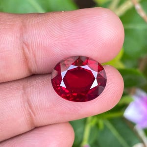 Könnte beinhalten: Ein tiefroter, oval geformter Edelstein mit Facettenschliff. Der Stein weist eine brillante, transparente Klarheit und einen satten karmesinroten Farbton auf, der das Licht durch seine präzise angeordneten geometrischen Facetten reflektiert.