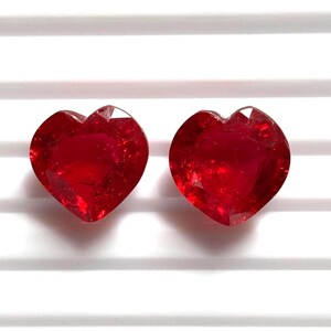 Könnte beinhalten: Zwei leuchtend rote, herzförmige Edelsteine mit einem Facettenschliff, der das Licht reflektiert. Diese losen Steine haben einen tiefen karmesinroten Farbton und eine polierte Oberfläche, die eine symmetrische Herzsilhouette mit scharfen Kanten zeigt.