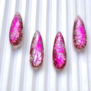 Könnte beinhalten: Vier tropfenförmige, facettierte Edelsteine in einer Reihe. Jeder klare Stein weist interne Einschlüsse von leuchtend neonpinken, nadelartigen Filamenten auf, die sich durch den transparenten Körper ziehen und ein helles, geometrisches Muster bilden.