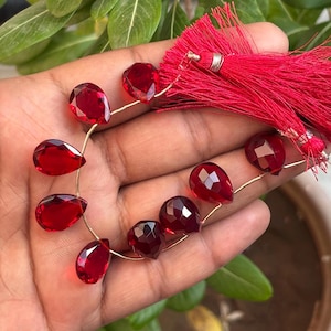 Könnte beinhalten: Eine Kette aus facettierten, tropfenförmigen und runden roten Edelsteinen. Die Edelsteine sind auf einem dünnen Golddraht aufgefädelt. Eine rote Quaste ist an der Kette befestigt. Die Edelsteine werden in einer Hand gehalten.
