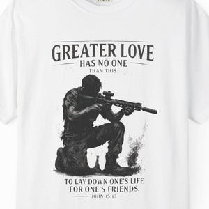 May include: White t-shirt featuring a black and white illustration of a person kneeling with a rifle. The shirt includes the text "GREATER LOVE HAS NO ONE THAN THIS" and "TO LAY DOWN ONE'S LIFE FOR ONE'S FRIENDS. JOHN 15:13."