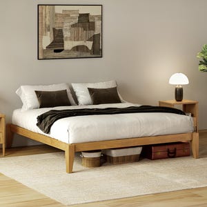 May include: A light wood bed frame with a white comforter and dark brown throw blanket. Two matching nightstands with lamps flank the bed. A framed abstract artwork hangs above the bed. Woven baskets and a suitcase are stored under the bed.