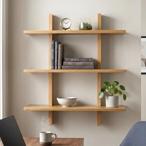 May include: A three-tiered light wood wall shelf featuring vertical supports. The shelves hold a round analog clock, a glass terrarium, three vertical books labeled DESIGN, three horizontal books labeled DESIGN, a white bowl, and a green plant in a pot.
