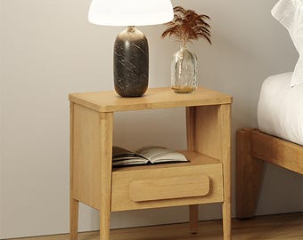 May include: A light-toned wooden nightstand featuring a single drawer with a rectangular handle and an open storage shelf. On top sits a lamp with a dark marble-patterned base and a white dome shade, alongside a clear glass vase with dried foliage.