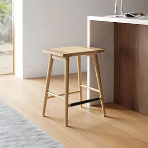 May include: A light wood bar stool with a woven seat and four angled legs. A black leather-like band is attached to the front legs. The stool is set against a light-colored wall and wooden floor.