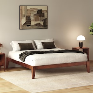 May include: A low-profile wooden bed frame with a white mattress, two white pillows, two dark brown rectangular accent pillows, and a dark brown knit throw blanket. The bed is flanked by two wooden nightstands, one featuring a lamp with a white dome shade.