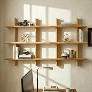 May include: A light-toned wooden wall-mounted shelving unit with three horizontal tiers and vertical dividers. The shelves hold vintage cameras, stacks of books, geometric wire sculptures, and ceramic vases, creating a structured display.