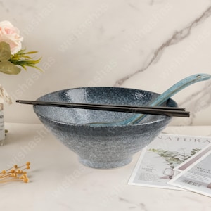 May include: A speckled blue ceramic ramen bowl with a matching blue ceramic spoon and black chopsticks. The bowl has a wide rim and a textured surface. The spoon has a curved handle. The chopsticks are straight and smooth.