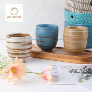 以下が含まれることがあります： 素朴なデザインのセラミックカップ3つ。1つは白地に茶色のストライプ、もう1つは青地に茶色の筆致、3つ目は茶色地に茶色のストライプです。カップは木製のトレイに置かれています。