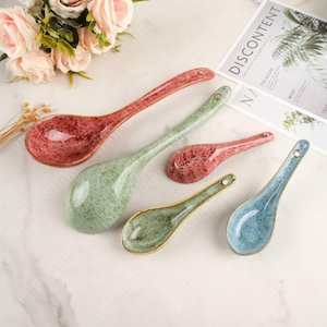 May include: A collection of five ceramic soup spoons in various sizes and colors. The spoons feature a speckled glaze in red, green, and blue hues, with a gold rim. Each spoon has a small hole at the end of the handle.