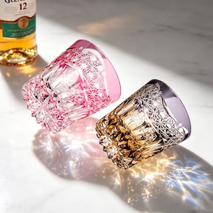 May include: Two crystal whiskey glasses featuring intricate geometric cut patterns. One glass is tinted in a vibrant pink hue, while the other displays a warm amber tone. Both glasses have a heavy, faceted base that creates light refractions.