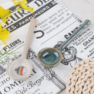 May include: Two ceramic spoons. One is beige with a cartoon fish design in the bowl. The other is green with a dark blue interior. The spoons are on a newspaper-style background with French text.