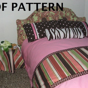 May include: A pink doll bed with a paisley headboard and a striped footboard. The bed has a pink blanket with a brown, green, and pink striped border. There are three pillows on the bed: one with brown polka dots, one with a zebra print, and one with a paisley print. A small side table with a striped cover sits next to the bed.