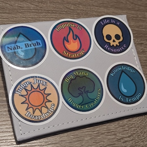 May include: A collection of six round stickers with various designs and text. The stickers are affixed to a gray, rectangular surface. The designs include a skull, a flame, a sun, a tree, and water droplets, each with accompanying text.