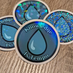 May include: Four round stickers with a blue water drop design and the text "Knowledge Is Tempo". The stickers have a holographic, iridescent finish, with a white border. The stickers are on a wooden surface.