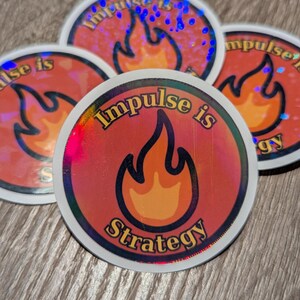 May include: A collection of round stickers with a holographic, iridescent finish. Each sticker features a stylized flame design in orange and yellow, with the words "Impulse is Strategy" in a bold, curved font. The stickers are on a wooden surface.