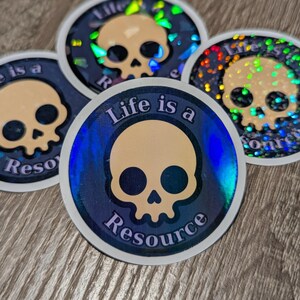 May include: A collection of circular stickers with a skull design. The stickers have a holographic effect with a blue and gold color scheme. The text "Life is a Resource" is printed on the stickers.