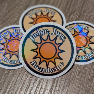 May include: Four round stickers with a holographic effect. Each sticker features the text "Judge, Jury, Boardwipe" in a circular design with a sun graphic in the center. The stickers have a white border and are on a wooden surface.
