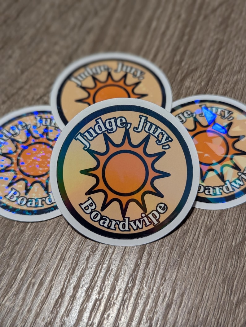 May include: Four round stickers with a holographic effect. Each sticker features the text "Judge, Jury, Boardwipe" in a circular design with a sun graphic in the center. The stickers have a white border and are on a wooden surface.