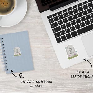 May include: A sticker with a tombstone design that reads "Here Lies My Energy." The sticker is shown on a notebook and a laptop, with arrows indicating its use as a notebook or laptop sticker. The tombstone is gray with green grass and pink and blue flowers.