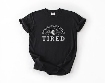 May include: Black t-shirt with the words "UNAPOLOGETICALLY TIRED" in white, arched above a crescent moon and stars design. The shirt is made of a soft material and has rolled-up sleeves.