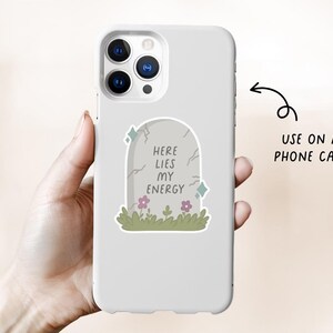 May include: A white phone case featuring a tombstone design with the text "HERE LIES MY ENERGY". The tombstone is gray with cracks, flowers, and green grass. The phone case is held in a hand.