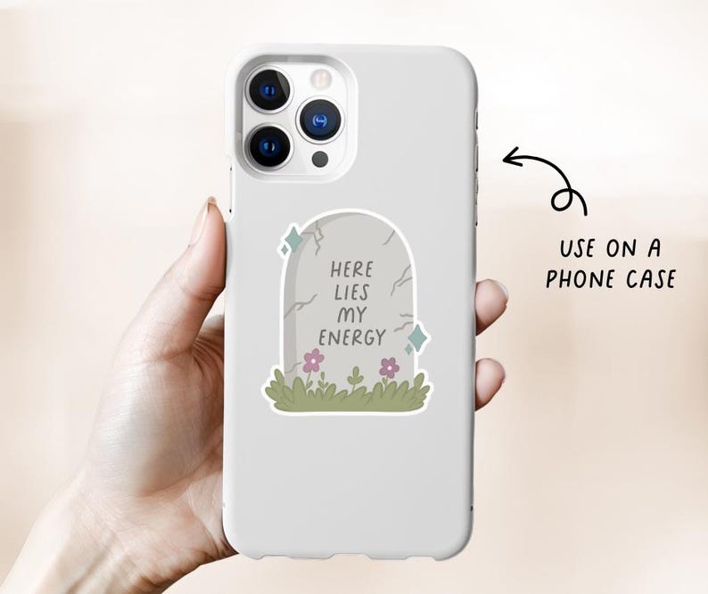 May include: A white phone case featuring a tombstone design with the text "HERE LIES MY ENERGY". The tombstone is gray with cracks, flowers, and green grass. The phone case is held in a hand.