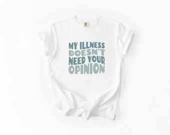May include: White t-shirt with the text "MY ILLNESS DOESN'T NEED YOUR OPINION" in shades of blue. The shirt has a crew neck and short sleeves. The text is stacked vertically.