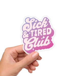 May include: A sticker with the text "Sick & Tired Club" in a retro font, with a gradient of pink and purple colors. The sticker has a white border and is held by a hand.