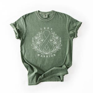 May include: A sage green t-shirt with rolled sleeves. The front displays a white floral design with a ribbon and the words "CRPS WARRIOR" in a circular arrangement. The shirt is made of a soft, comfortable material.