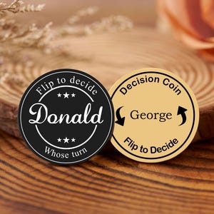 May include: Two decision coins, one black and one tan, with text. The black coin says "Flip to decide Donald Whose turn" in white. The tan coin says "Decision Coin George Flip to Decide" in black. The coins are on a wooden surface.