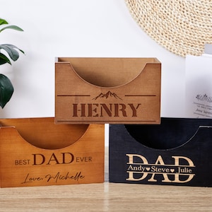 Könnte beinhalten: Drei Schreibtisch-Organizer aus Holz mit gebogenen Frontöffnungen. Einer ist mit "HENRY" und einer Berg-Grafik graviert. Ein anderer sagt "BEST DAD EVER, Love Michelle." Der dritte ist schwarz und trägt die Aufschrift "DAD" mit Namen.