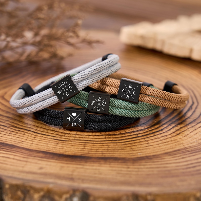 May include: Five rope bracelets in various colors: gray, black, green, and brown. Each bracelet features a black square clasp with engraved letters and numbers. The bracelets are displayed on a wooden surface.
