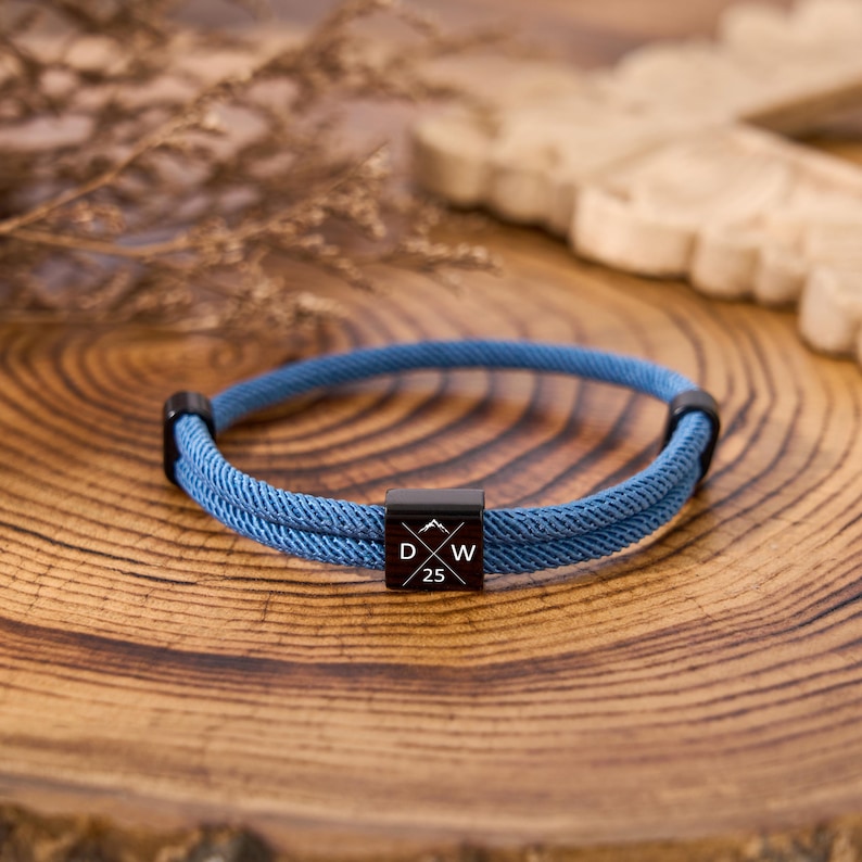 May include: Blue rope bracelet with black accents and a black square centerpiece. The centerpiece features a mountain graphic and the letters "D" and "W" with the number "25". The bracelet is displayed on a wooden surface.