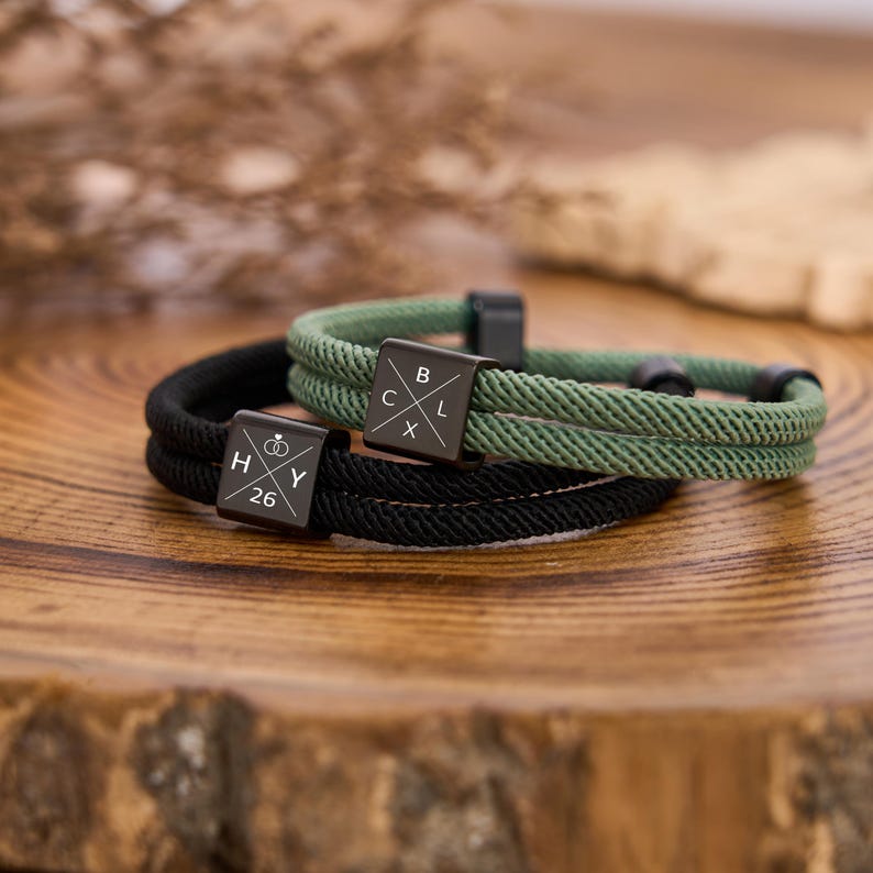 May include: Two rope bracelets, one black and one green, with black metal square clasps. The black bracelet has the letters "H", "Y", and the number "26" on its clasp, along with a ring symbol. The green bracelet has the letters "B", "C", "L", and "X" on its clasp.