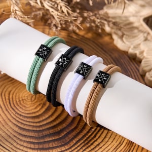 May include: Four rope bracelets in green, black, white, and tan, each with a black square charm. The charms have white lettering and symbols. The bracelets are displayed on a white cylinder against a wooden background.