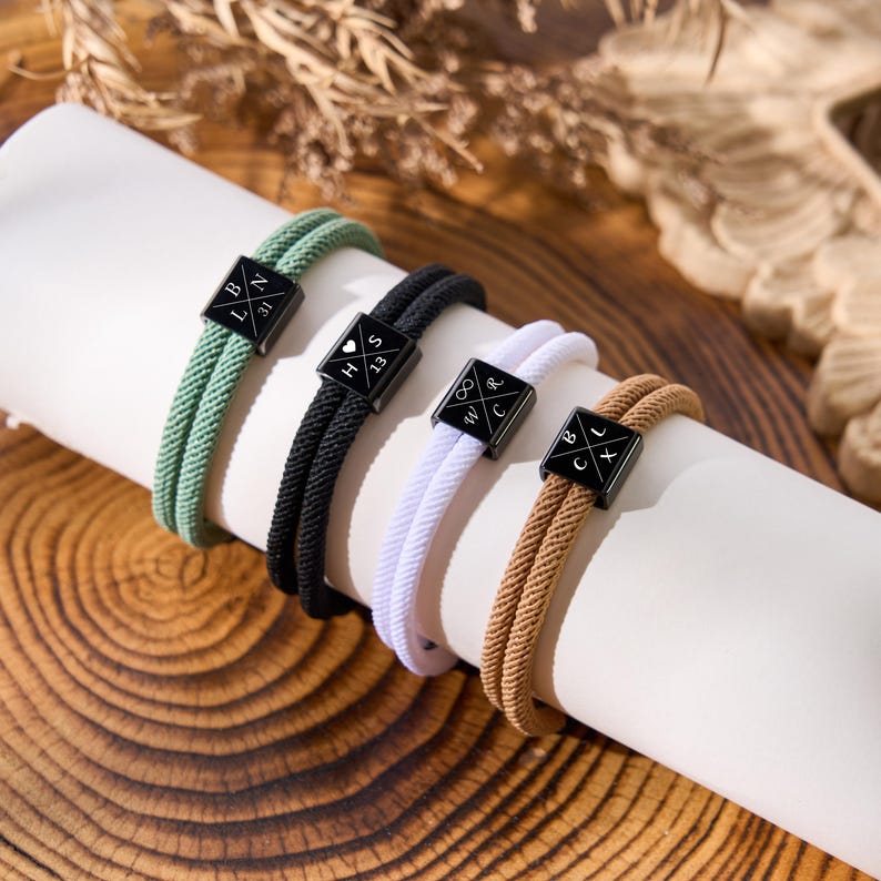 May include: Four rope bracelets in green, black, white, and tan, each with a black square charm. The charms have white lettering and symbols. The bracelets are displayed on a white cylinder against a wooden background.