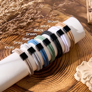 May include: A display of several bracelets in various colors, including khaki, gray, blue, green, black, white, and brown. Each bracelet features a woven design and a black square clasp. The colors are labeled with text.