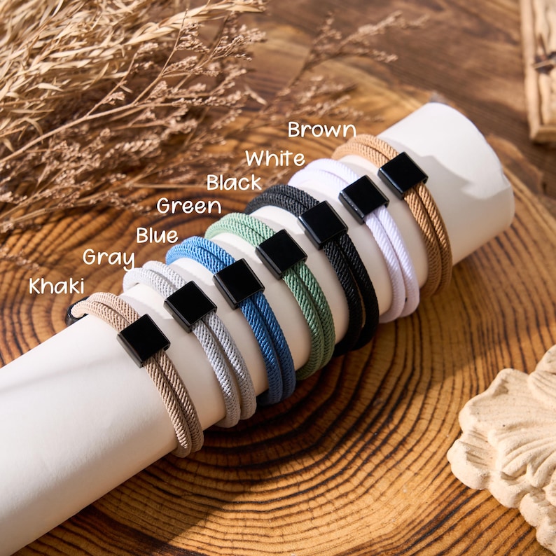 May include: A display of several bracelets in various colors, including khaki, gray, blue, green, black, white, and brown. Each bracelet features a woven design and a black square clasp. The colors are labeled with text.