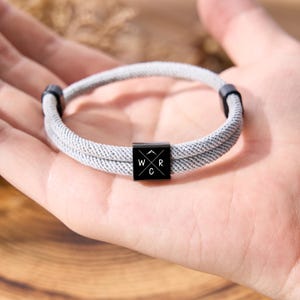May include: A gray rope bracelet with a black square centerpiece. The centerpiece has the letters "W", "R", and "C" with a mountain graphic. The bracelet is held in a hand.