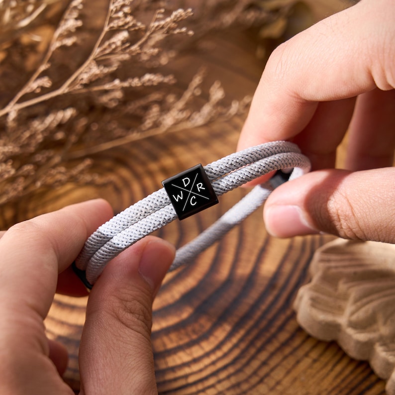 May include: A gray rope bracelet with a black square clasp. The clasp has the letters "D", "R", "W", and "C" in white. The bracelet is held by hands against a wooden background.