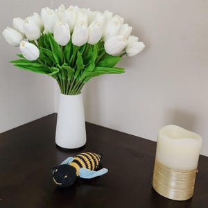 May include: A white vase holds a bouquet of white tulips with green leaves. A crocheted bee with black, yellow, and blue details rests on a dark surface, next to a cream-colored candle with a gold band.