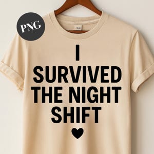 May include: A beige t-shirt with rolled-up sleeves. The shirt features the text "I SURVIVED THE NIGHT SHIFT" in bold black letters, with a black heart symbol below the text. The shirt is hanging on a wooden hanger.
