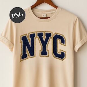 May include: A cream-coloured t-shirt with rolled-up sleeves. The shirt features the letters "NYC" in a navy blue, textured font with a gold glitter outline. The shirt is hanging on a wooden hanger.